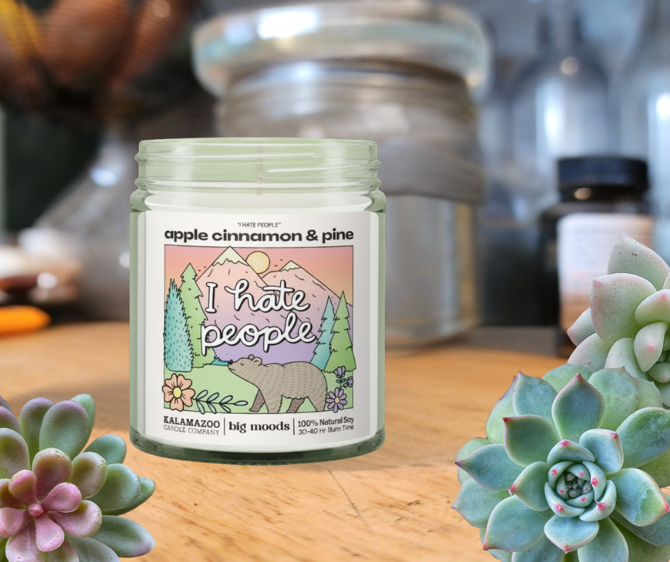Candle jar labeled 'I hate people' with decorative design on a wooden surface with succulents.