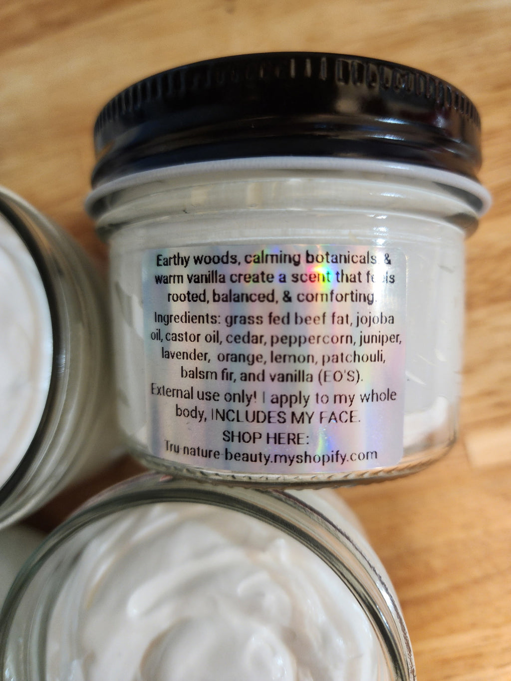 A close-up of a clear glass jar with a black lid containing white cream, showing the product label text on a wooden surface. Other partial jars are visible at the edges.