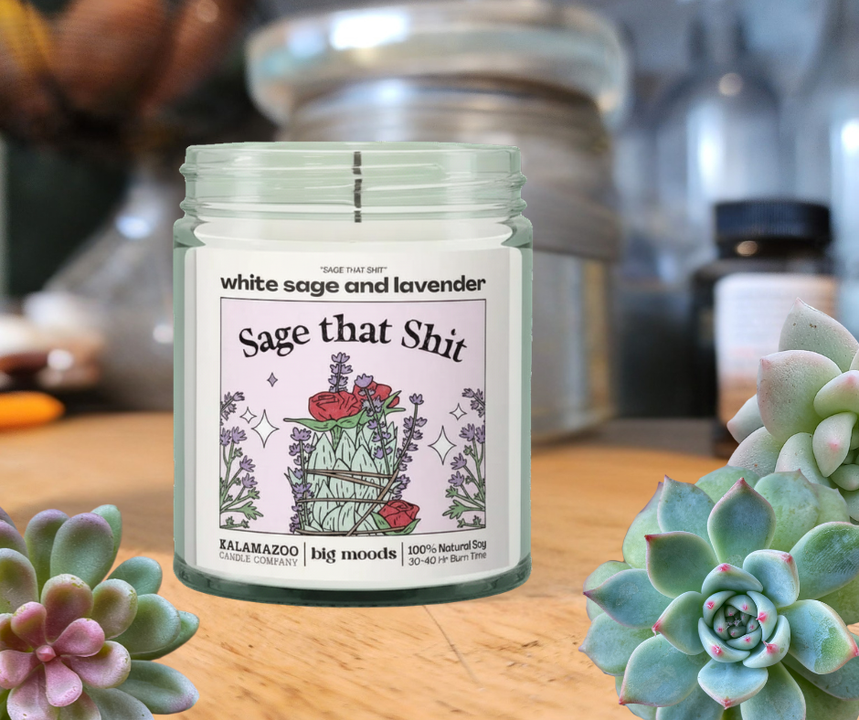 Candle jar labeled 'Sage that Shit' with succulents on a wooden surface