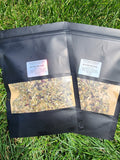 Two black resealable stand-up pouches of Roofted Revival loose-leaf tea placed on green grass. Each pouch has a clear window showing the dried tea blend inside and a white label near the top.