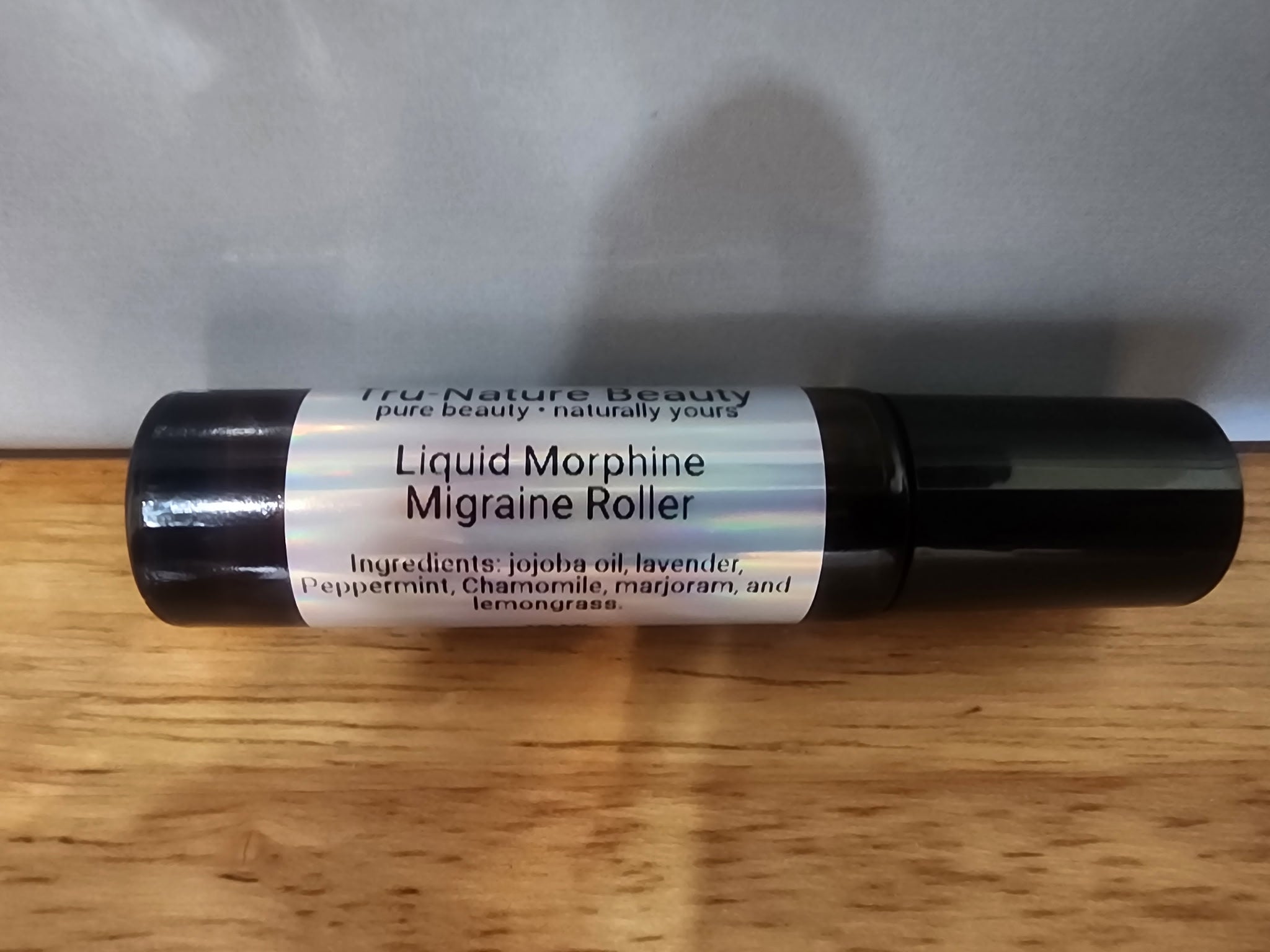 A black cylindrical roller bottle labeled 'Liquid Morphine Migraine Roller' by True Nature Beauty, resting on a wooden surface against a gray background.