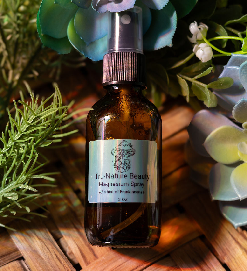 Brown glass bottle of Tru-Nature Beauty Magnesium Spray with a label, surrounded by greenery on a wooden surface.