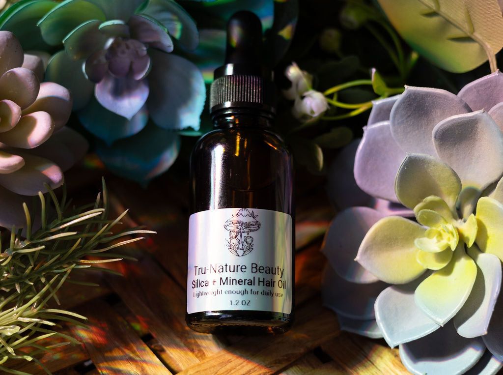 Bottle of Tru Nature Beauty Silica + Mineral Hair Oil surrounded by succulent plants on a wooden surface.
