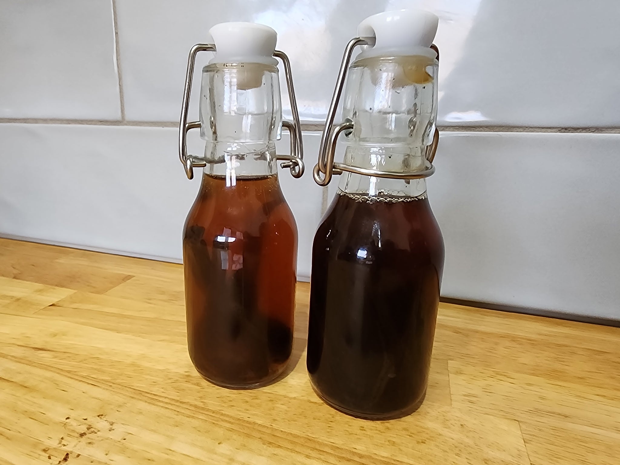 Two swing-top glass bottles with white closures, one containing amber liquid and the other a very dark brown liquid, set on a wooden surface against a white tiled wall.