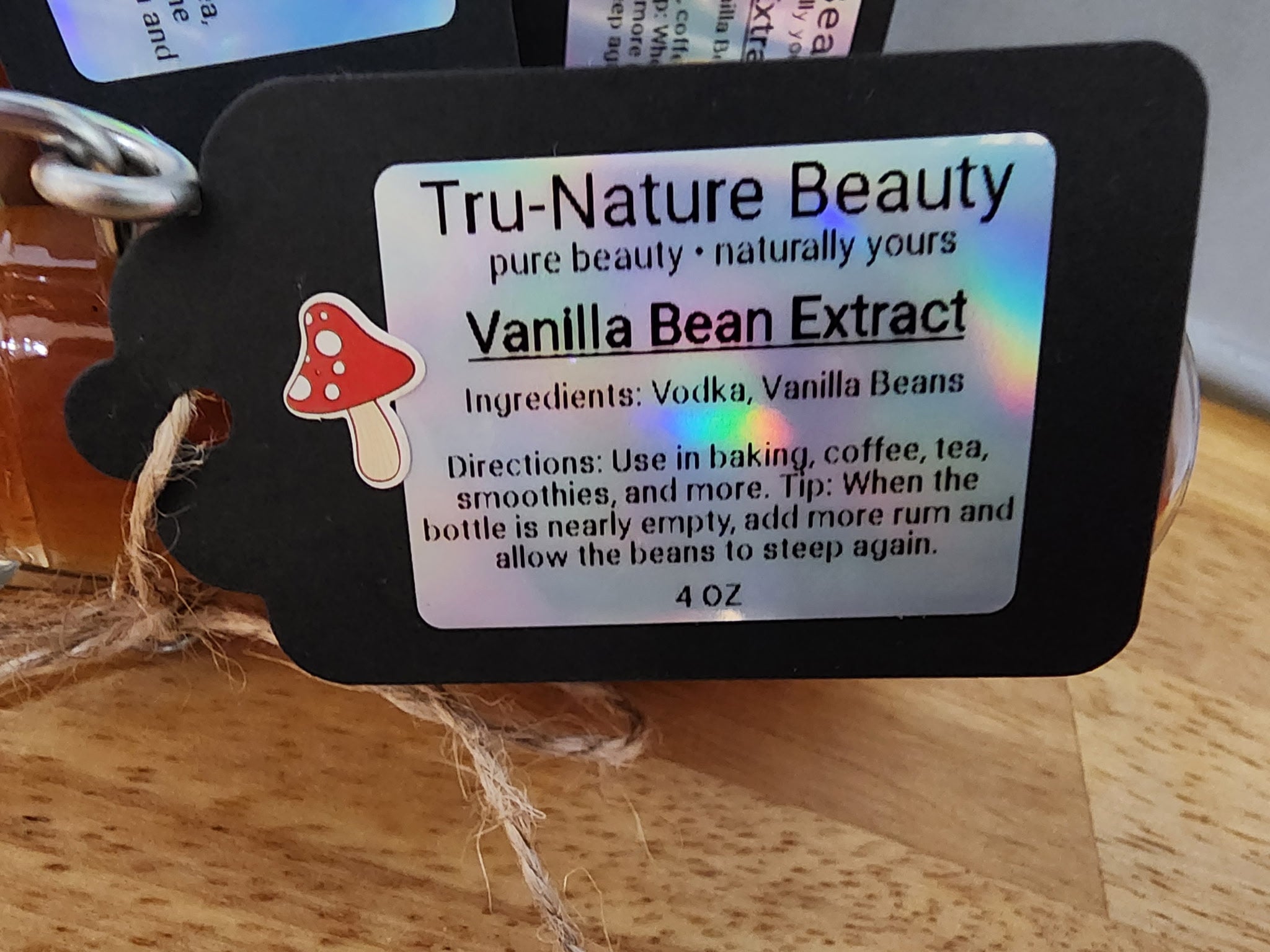 A close-up of a black product tag labeled 'Tru-Nature Beauty Vanilla Bean Extract' with a small red-and-white mushroom icon, photographed on a wooden surface.