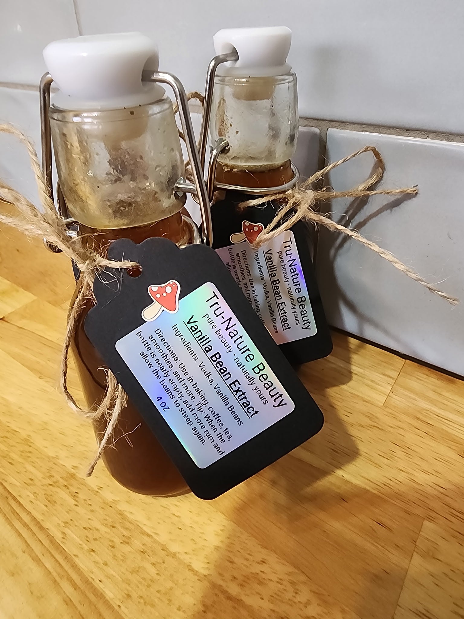 Two small glass jars filled with vanilla bean extract, tied with twine and featuring black product tags that read 'The Artful Beauty Vanilla Bean Extract,' set on a wooden surface against a white tiled background.