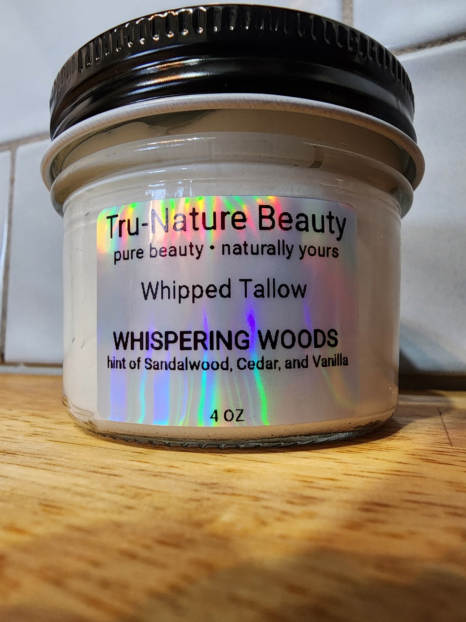 A 4 oz jar of Tru-Nature Beauty Whipped Tallow body butter in the scent Whispering Woods, resting on a wooden surface with a tiled background.